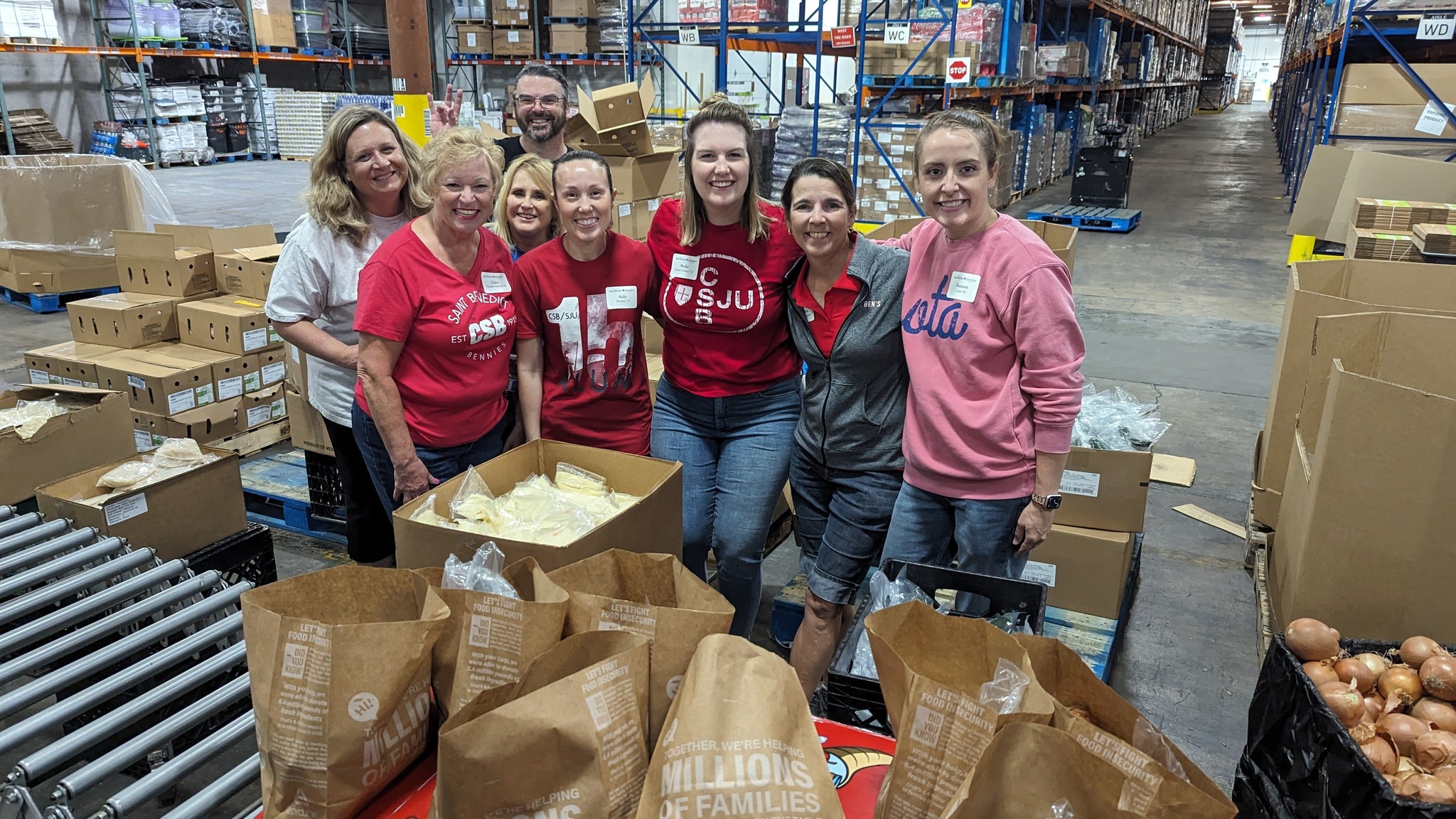 A group of eight people, wearing casual clothing with some in red shirts, stand smiling in a warehouse among boxes and bags. They appear to be volunteering, surrounded by shelves with supplies and food items.