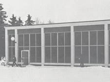 Black and white photo of a building with large windows and vertical columns. Snow covers the ground. Two individuals, one pushing a bicycle and the other walking, are near the building. Pine trees are visible in the background.