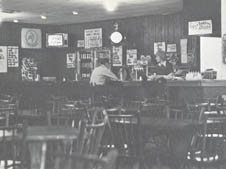 A black-and-white photo of a nearly empty bar with a few people seated at the counter. The bar is lined with bottles, and there's a clock on the wall. Several chairs and tables fill the foreground. The room has a vintage feel, with posters and signs on the walls.