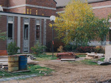 A construction site with scattered materials, metal drums, wooden planks, and other debris on the ground. A brick building with windows under renovation is in the background, and a leafy tree with yellow foliage stands to the right.