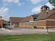 A modern, single-story, red-brick building with a gabled roof and several windows. It has a small courtyard and is surrounded by a manicured lawn. A streetlamp is visible on the right, and the sky is partly cloudy.