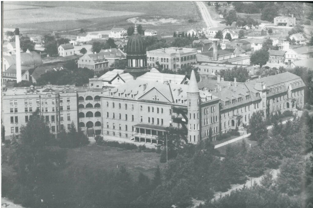 A black-and-white aerial photograph of a large, historic building complex featuring multiple connected structures with varied architectural styles. The complex is surrounded by trees and situated in a town with several houses and smaller buildings visible in the background.