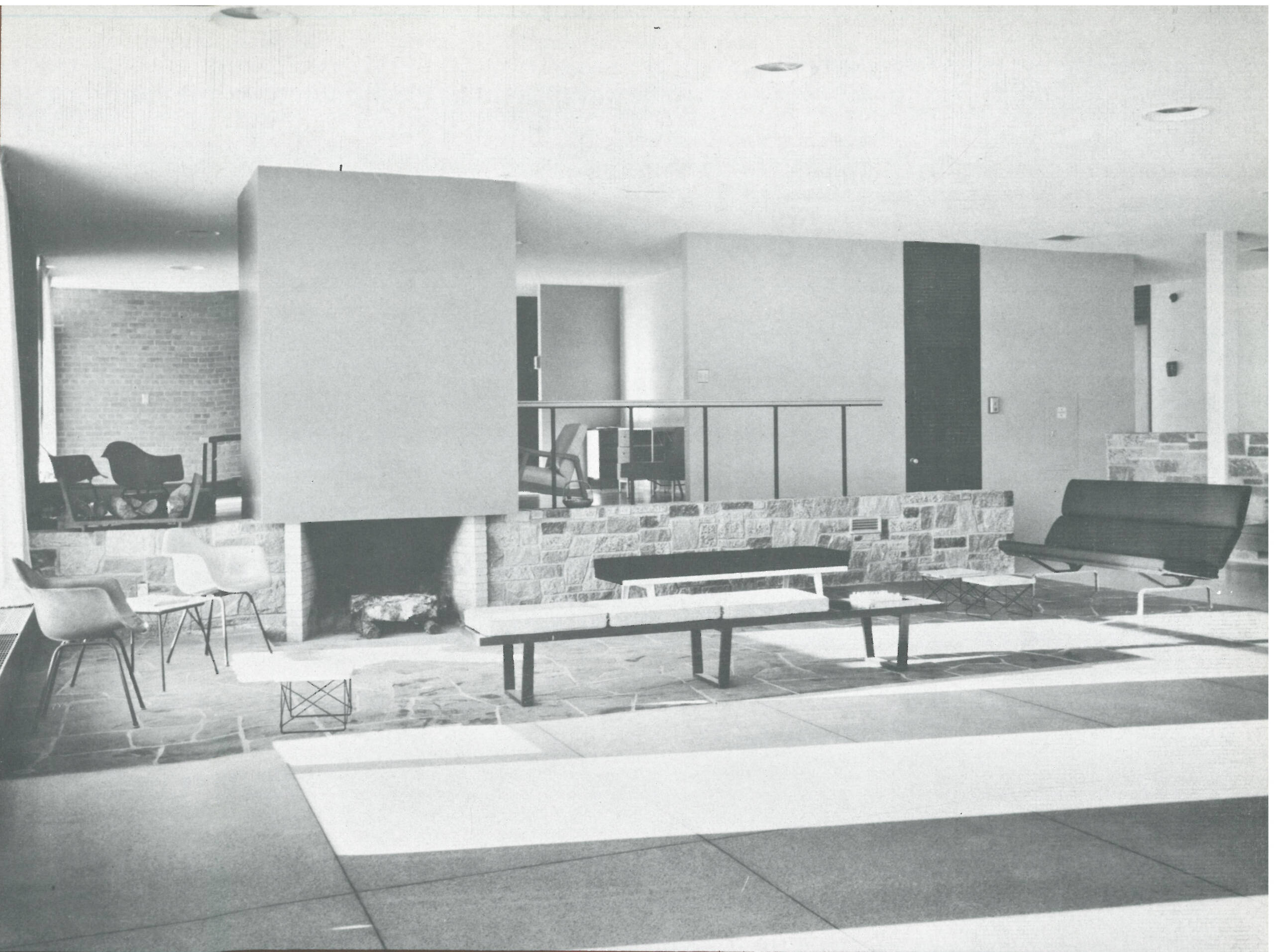 Black and white photo of a modern, open-concept interior living space. It features a minimalist design with a stone fireplace, low benches, Eames chairs, and large windows letting in natural light. The room is defined by clean lines and uncluttered surfaces.