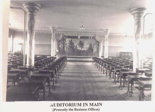 A vintage photograph of an auditorium with rows of wooden desks and chairs facing a stage. Ornate columns and a mural with a nature scene decorate the room. The caption reads: "AUDITORIUM IN MAIN (Presently the Business Offices).