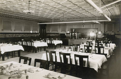 A vintage dining room with a high decorated ceiling, empty tables covered with white tablecloths, and wooden chairs. The room features a wooden buffet area at the back and is lit by hanging lights. Elegant table settings are ready for guests.