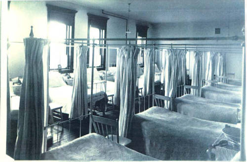 A sepia-toned photograph of a large hospital ward, featuring rows of beds separated by curtains. The room has a high ceiling and several windows, allowing natural light to filter in. Simple wooden chairs are placed next to each bed.