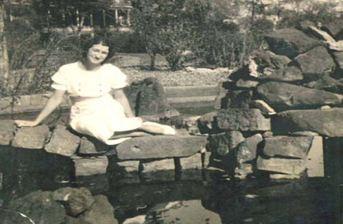 A woman in a white dress sits on a stone wall by a pond, smiling. The background features trees and a wooden structure, suggesting a garden or park setting. The photo has a vintage, sepia tone.