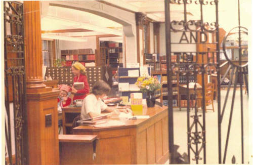 A library scene with a woman sitting at a wooden desk, writing. Bookshelves filled with books are in the background. Another person in a red outfit and yellow hat stands nearby, holding books. The foreground has ornate metalwork.