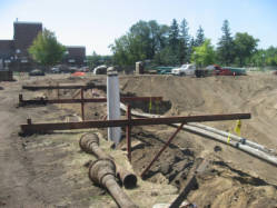 Construction site with large pipes laid in a dirt trench. Metal beams are used for support across the trench. Buildings and vehicles are visible in the background, with trees lining the horizon under a clear sky.