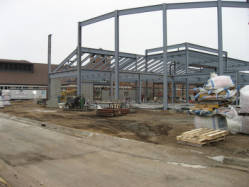 A construction site with partially built metal framework for a new building. The background shows existing structures, and the ground is covered with dirt and construction materials. Skids and wrapped items are visible in the foreground.