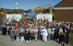 A large group of people gathered outdoors in front of construction equipment and banners. They are posing for a group photo on a sunny day. The scene suggests a community event or celebration.