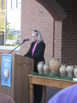 A woman is speaking at a podium with a microphone, wearing a black jacket and pink shirt. Several clay pots are displayed on a table nearby. A poster titled "Our Place in the World" is attached to the podium. A brick wall and window are in the background.