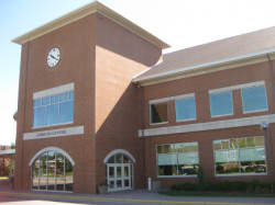 A two-story brick building with a large clock tower. The structure has several windows and an entrance with glass doors. The sign reads "Library and Technology Center." The surrounding area has a small amount of greenery.