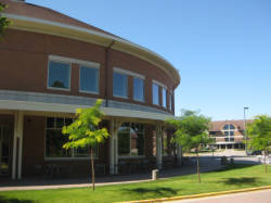 A curved brick building with large windows stands under a clear blue sky. Young trees line the sidewalk, and another building is visible in the background.