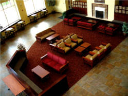 A spacious lounge area with several couches and armchairs arranged on a red carpet. The room features large windows, wooden tables, and a fireplace, creating a cozy and inviting atmosphere.