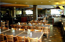 A spacious, empty restaurant with wooden tables and chairs neatly arranged. The lighting is warm, and there are cushioned booths along the back. Large windows in the background let in natural light.