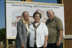 Three people, two women and a man, stand smiling in front of a sign for the Gorecki Dining and Conference Center. The sign includes an illustration of the building. The people are dressed in casual and business attire.