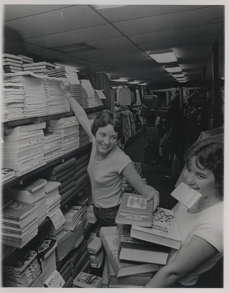 Two women in a bookstore smile as they organize piles of books. One reaches for a book on a high shelf, while the other holds several books and has a paper in her mouth. Shelves filled with various books line the narrow aisle.