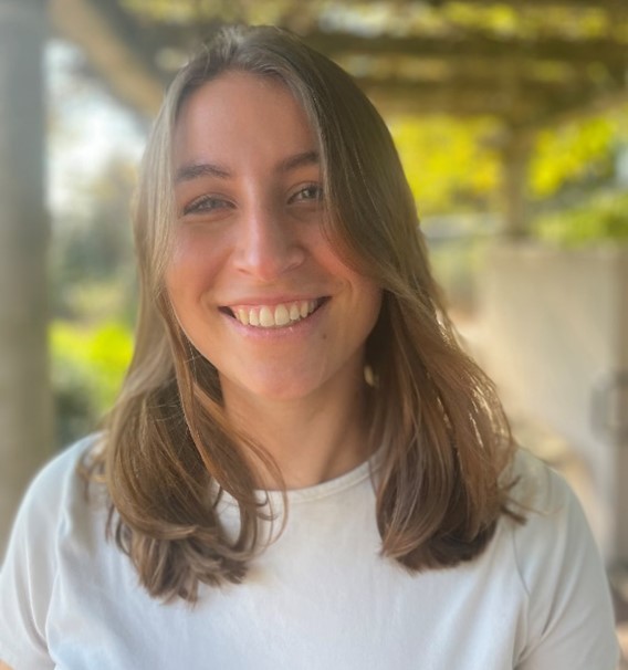 A person with shoulder-length light brown hair and a white shirt is smiling in an outdoor setting with greenery and a pathway in the background. The image is bright and sunny.