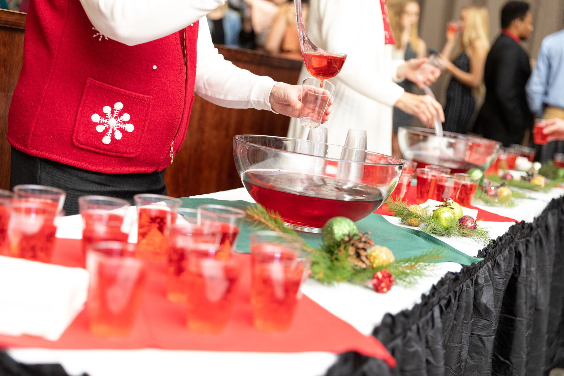 People in festive attire serving red punch at a decorated table with holiday ornaments and a black table skirt. In the background, other guests mingle in a festive setting.