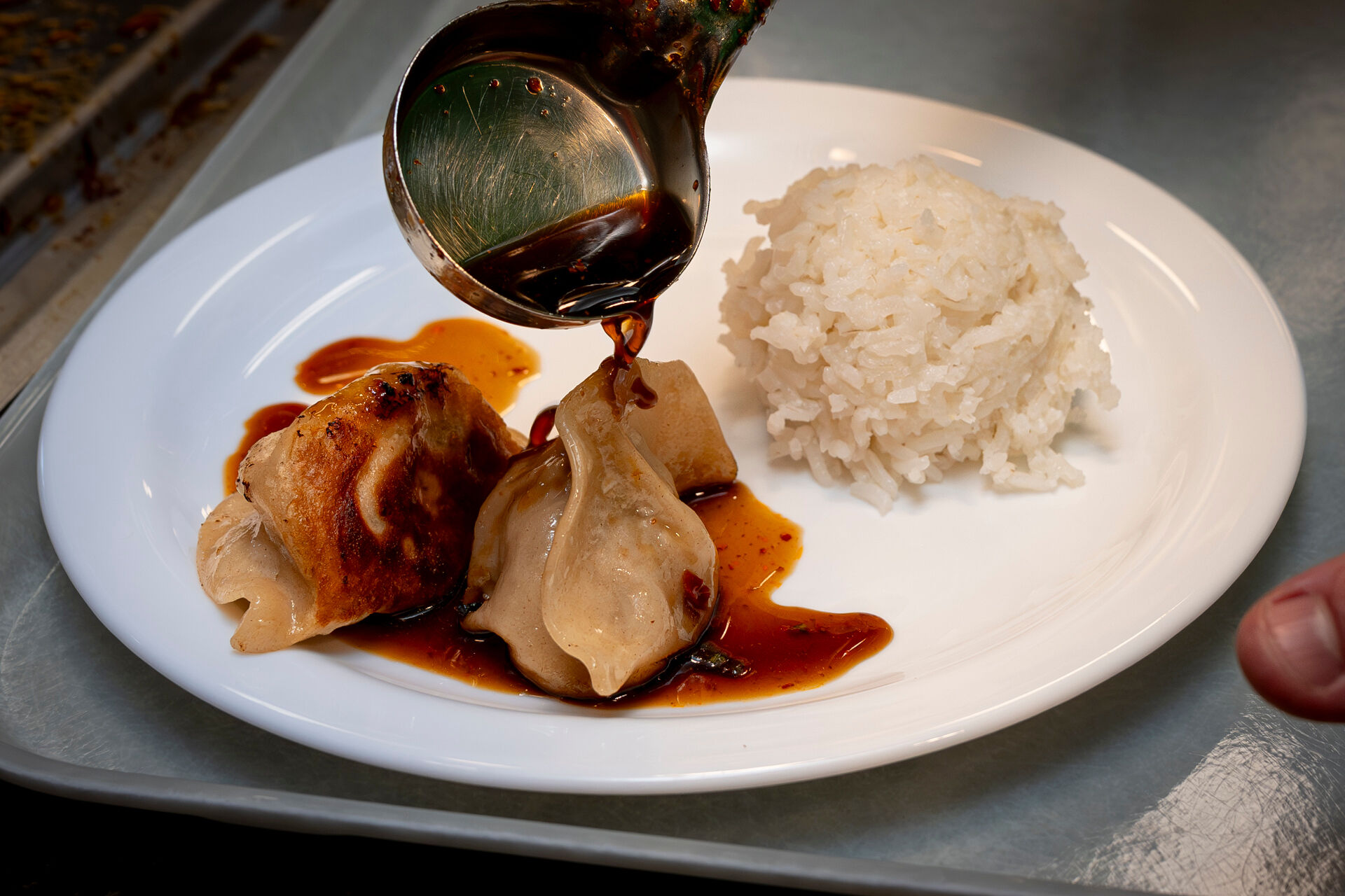 A plate with two dumplings drizzled with a dark sauce from a ladle, accompanied by a serving of white rice on the side.