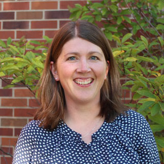 A woman with shoulder-length brown hair, wearing a navy blue blouse with a white dot pattern, stands smiling in front of a brick wall and lush green foliage.