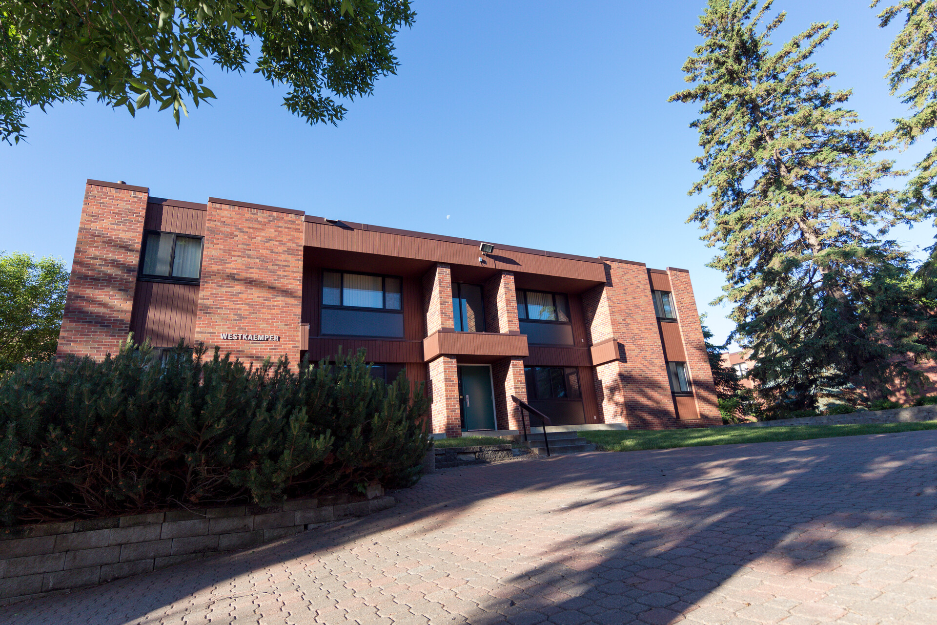 A modern, two-story brick building with large windows and a sign reading "ASBURY CAMPUS" stands amid green trees. A paved walkway leads to the entrance, and the sky is clear and blue.