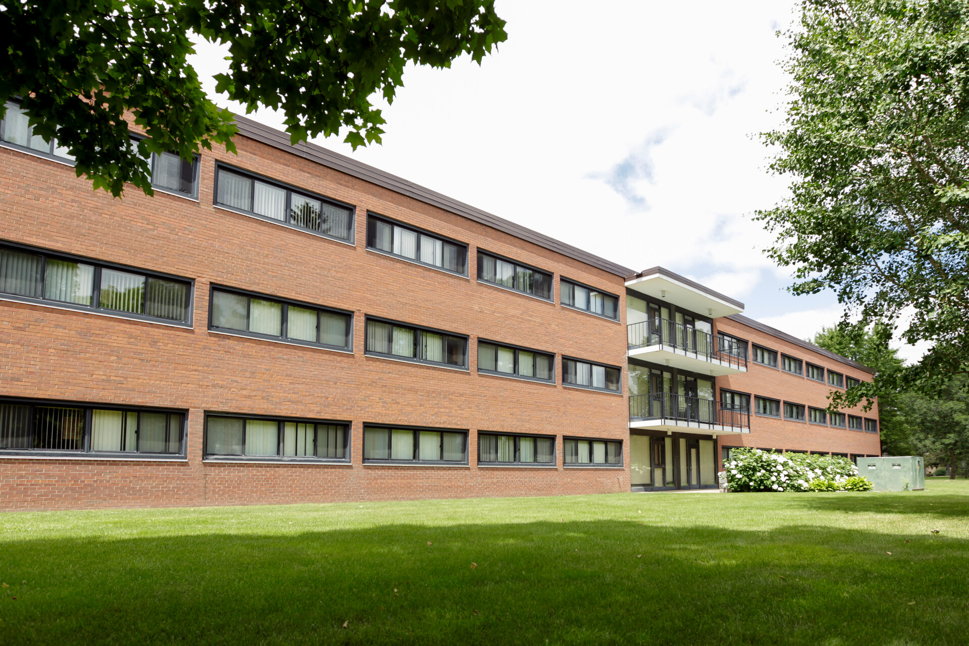 A three-story brick building with numerous rectangular windows. There is a modern glass entrance with balconies on the second and third floors. The building is surrounded by a well-maintained lawn and trees. The sky above is partly cloudy.