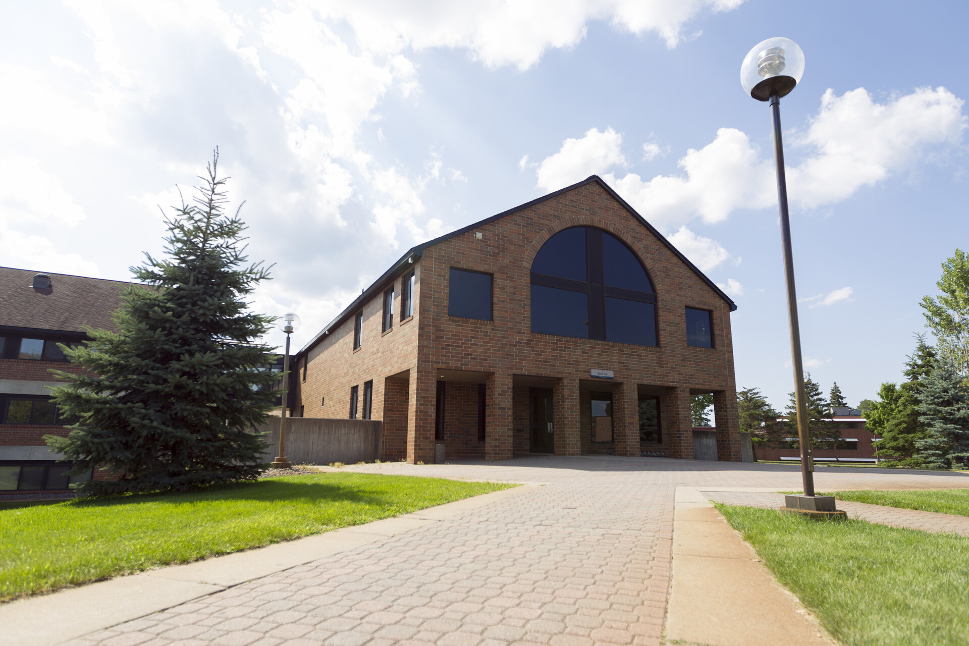 A two-story brick building with large, dark windows and an arched roof section stands under a partly cloudy sky. A pathway leads to the entrance, lined with grass, a streetlamp, and an evergreen tree to the left of the building.