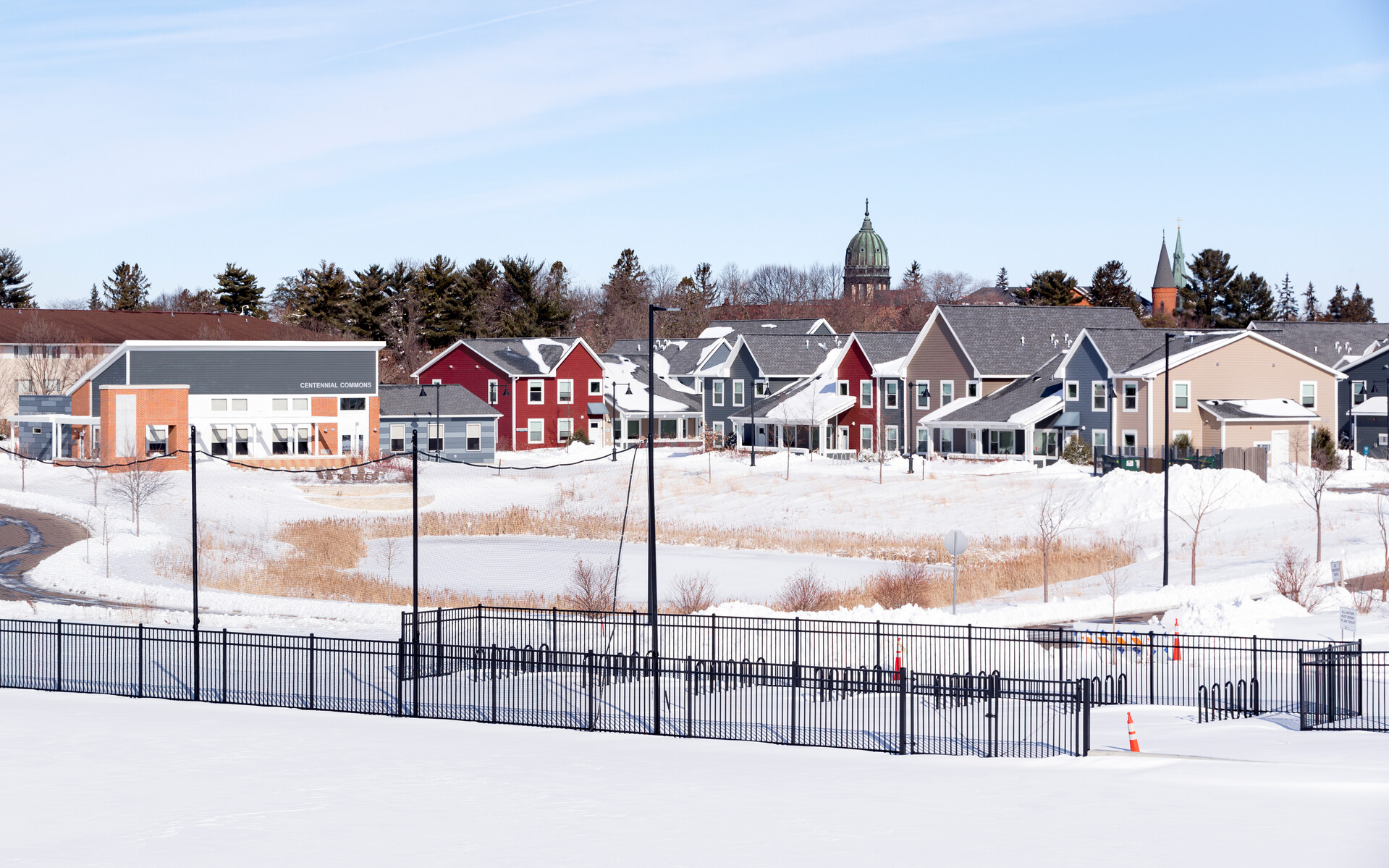 A snowy landscape featuring a suburban neighborhood with colorful houses, including red, beige, gray, and white homes. Snow covers the ground, and a pond is partially visible behind a black fence. Trees and a church with a dome are seen in the background.