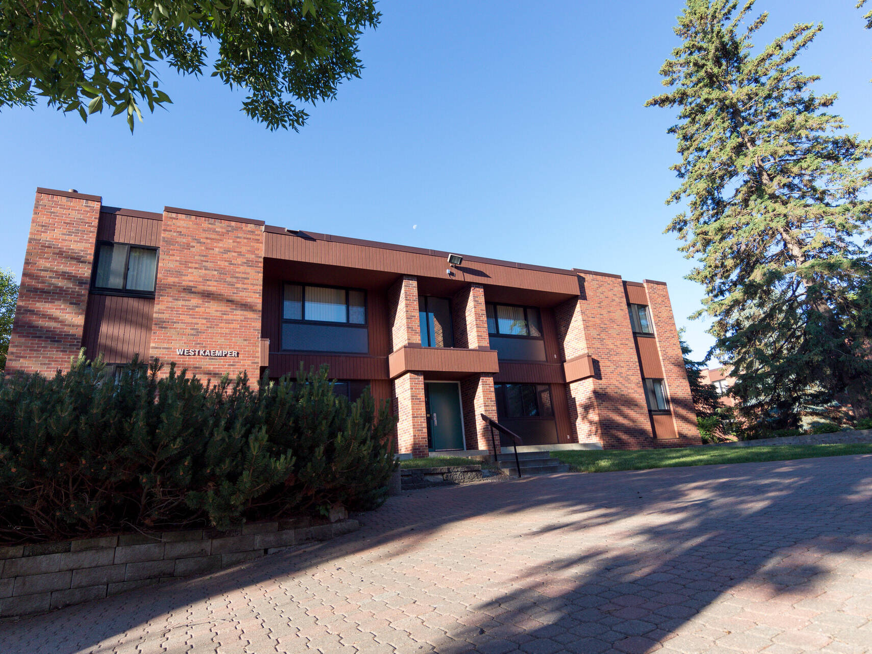 A modern, two-story brick building with large windows and a sign reading "ASBURY CAMPUS" stands amid green trees. A paved walkway leads to the entrance, and the sky is clear and blue.