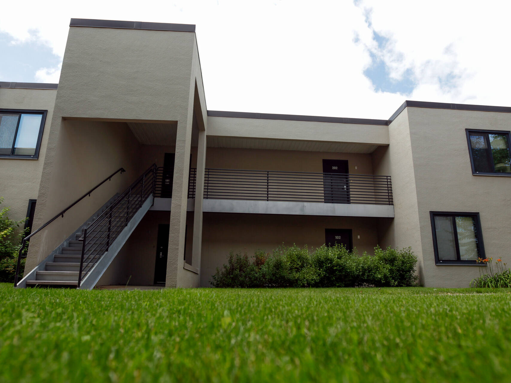 A two-story beige building with a modern design. It features a central staircase leading to the upper floor. The foreground shows a well-maintained green lawn with a few shrubs near the building. The sky is partly cloudy.