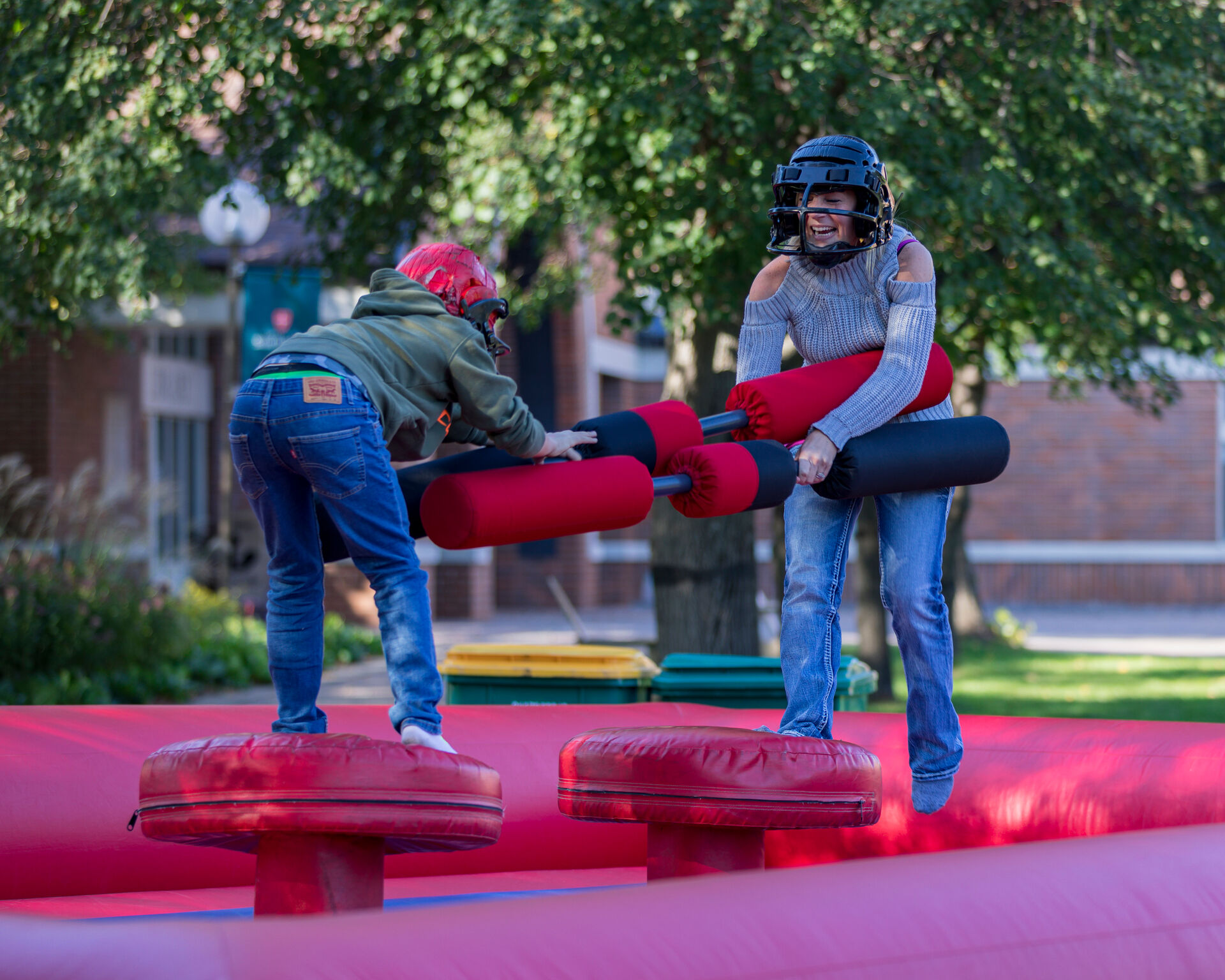 Two people wearing helmets and protective gear engage in a friendly jousting match on an inflatable arena. They balance on red platforms while holding padded jousting sticks. The background shows a tree and a building.