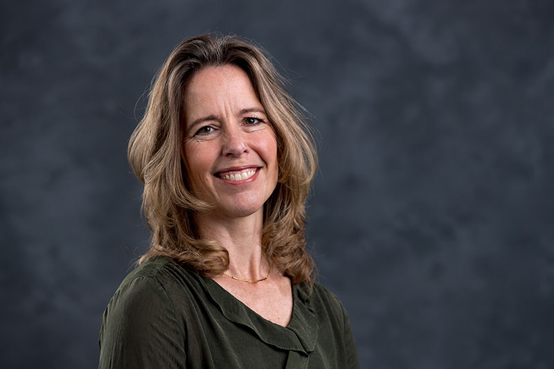 A woman with shoulder-length sandy brown hair smiles at the camera. She is wearing a dark green blouse and a delicate necklace. The background is a mottled gray-blue.