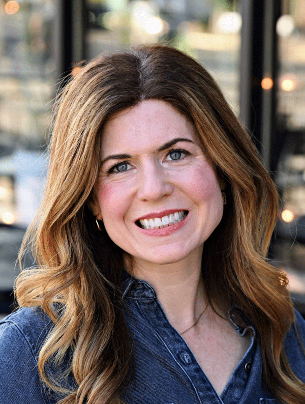 Madey Israelson, Associate Professor and Director of Assessment. A smiling woman with long wavy brown hair wearing a denim shirt. She is standing outdoors with blurred lights and a reflective background.