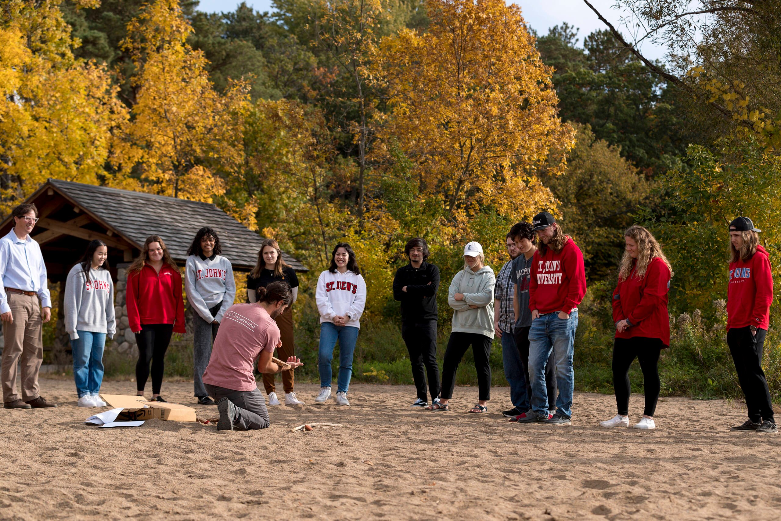 A group of people stands on a beach, facing a person kneeling on the ground who appears to be demonstrating something. The group is diverse and casually dressed, with colorful trees and a wooden shelter visible in the background.