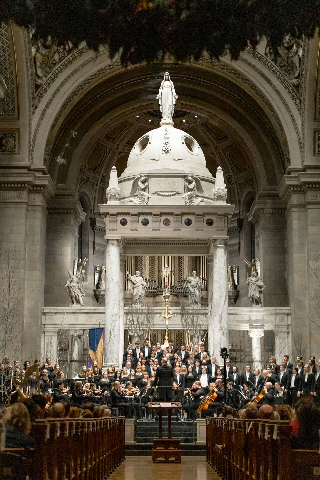 A choir and orchestra perform in a grand church. The conductor stands in front, directing musicians seated below an ornate altar with statues and a large dome. The space is richly decorated, with a central sculpture of a woman atop the dome.