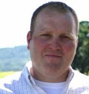 A man with short hair is standing outdoors. He is wearing a light-colored, collared shirt, and the background shows a blurred scenic view with greenery and hills under a clear sky.