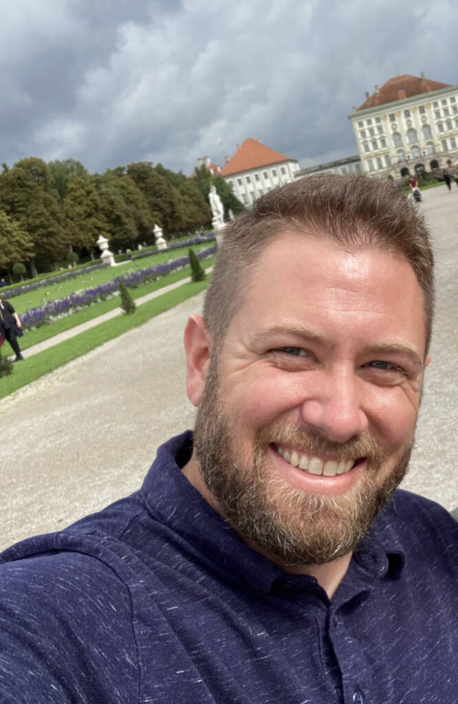 A man with a beard and short hair smiles at the camera while taking a selfie in front of a manicured garden with purple flowers and a historic building. The sky is partly cloudy, and there are trees and other people in the background.