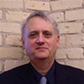 A middle-aged man with short gray hair is wearing a dark suit and tie. He stands against a backdrop of an exposed brick wall and has a neutral expression.