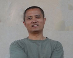 A person with short hair wearing a green shirt stands with arms crossed against a plain light-colored wall, smiling slightly.