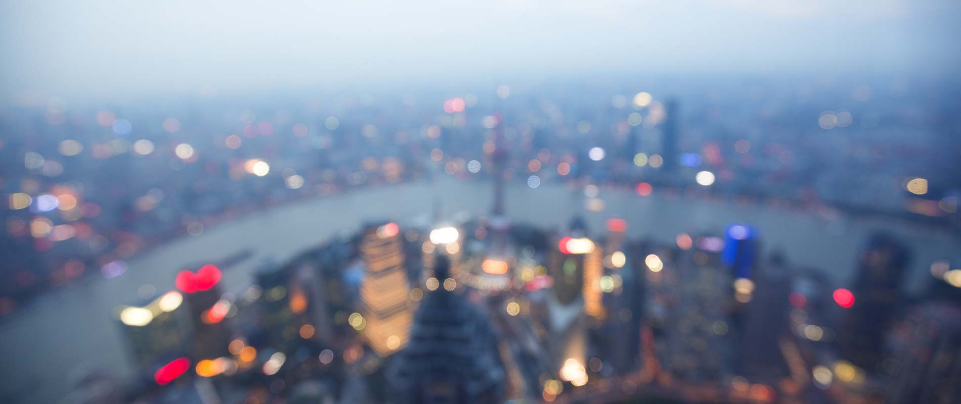 A cityscape at dusk with a river running through it. The image is blurred, causing the city lights on the buildings and streets to create a soft, out-of-focus glow.