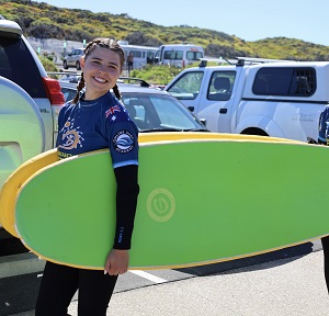 A person holding two surf boards in front of parked cars.