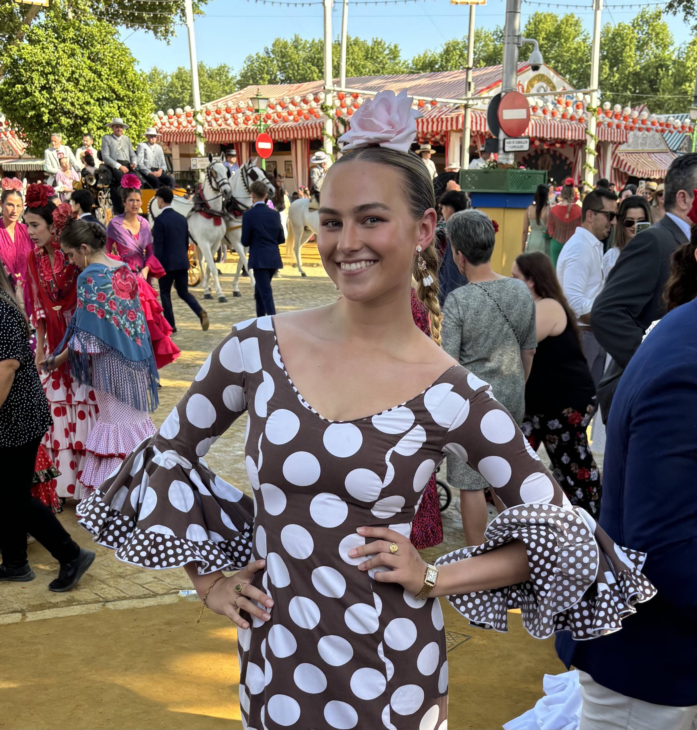 A woman dressed in a brown flamenco dress with white polka dots and ruffled sleeves stands smiling with her hands on her hips. She is wearing a pink flower in her hair, and in the background, people are dressed in traditional attire with decorated stalls and horses present.