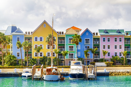 A row of colorful buildings, painted in pastel yellow, pink, blue, and other hues, lines a waterfront. Palm trees are scattered along the promenade, and several boats are docked in the clear turquoise water under a partly cloudy sky.