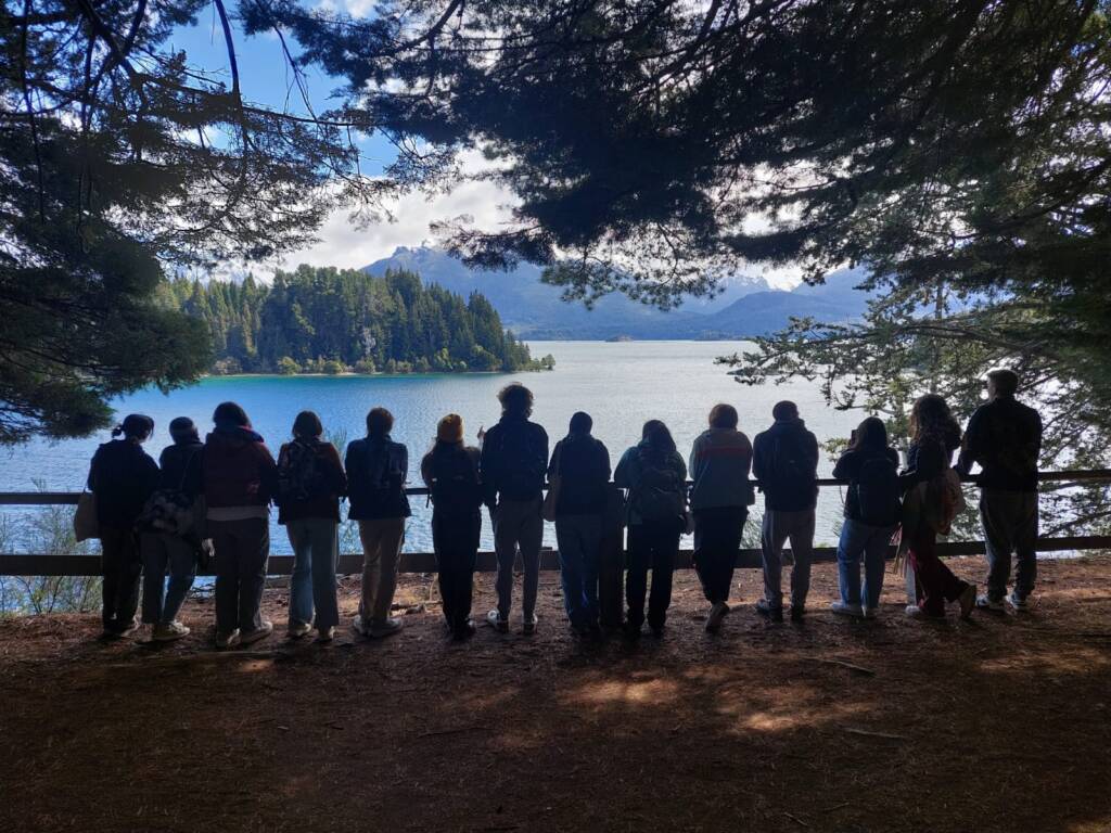 A group of people stand together facing a scenic view of a lake surrounded by trees. They are silhouetted against the water, and sunlight filters through the branches overhead, creating a peaceful, natural setting.