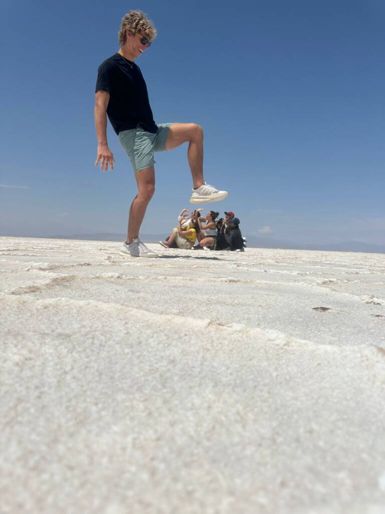 A person playfully poses on a salt flat, appearing to step on a group of people sitting further back. The sky is clear and blue, enhancing the playful illusion of the photo.