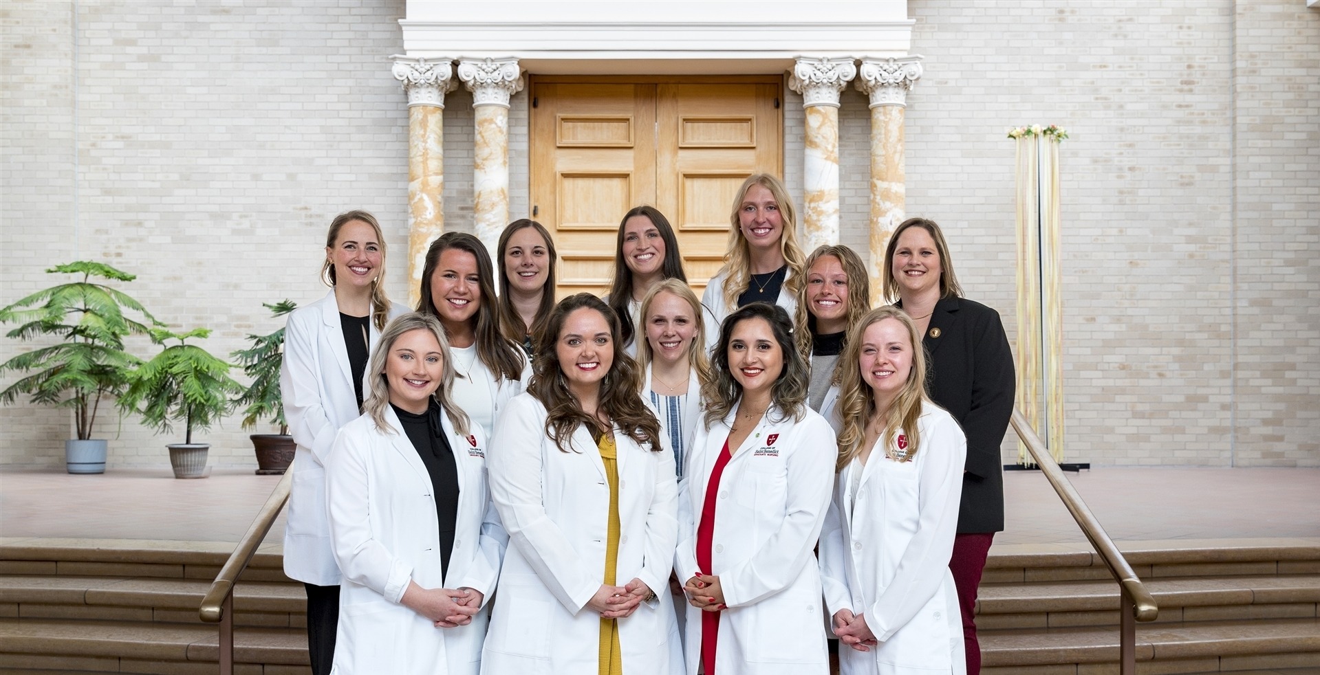 A group of thirteen women, some in white lab coats, are standing on steps inside a building with beige walls and columns. The women are smiling and posing for the camera. There are large wooden doors in the background.