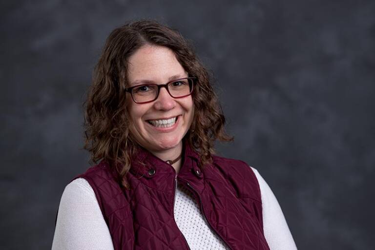 A smiling person with curly hair and glasses is wearing a maroon vest over a white shirt. The background is a textured dark gray.