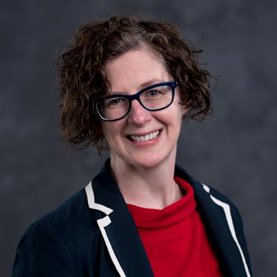 A person with curly, short brown hair, wearing glasses, a navy blazer with white trim, and a red top, smiles against a dark gray background.